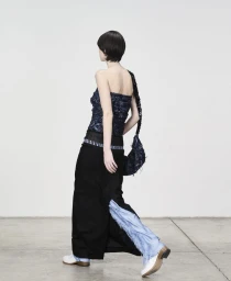 Model with dark hair walking away wearing a dark textured top, long black skirt, and white shoes against a plain wall.