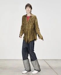 Model wearing an olive frayed button-down shirt and dark cuffed trousers standing against a plain white wall