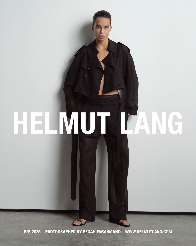 Model wearing a black cropped trench coat and matching trousers standing against a pale grey studio wall