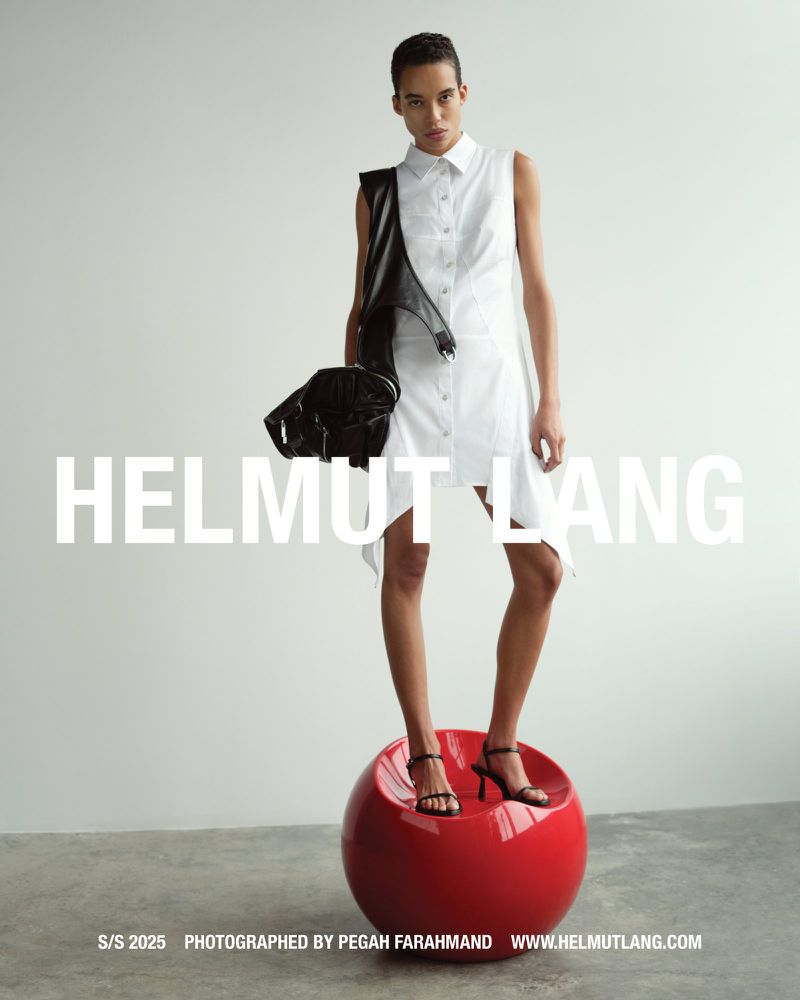 Model wearing white sleeveless shirt dress standing on a red sphere against a plain background