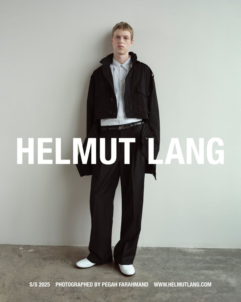 Full-length portrait of a male model wearing a black cropped jacket, white shirt, and black trousers against a neutral grey wall.