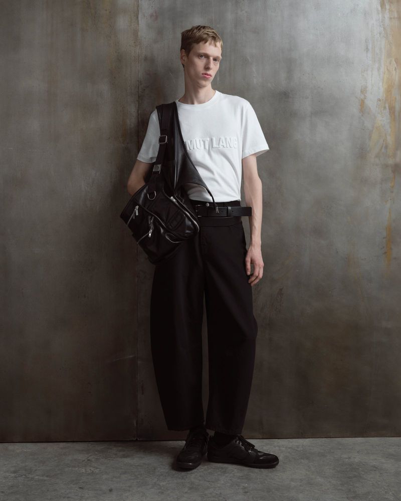 Full-length studio shot: a slender man with fair hair, in a white t-shirt, black wide-leg pants, and black leather bag.