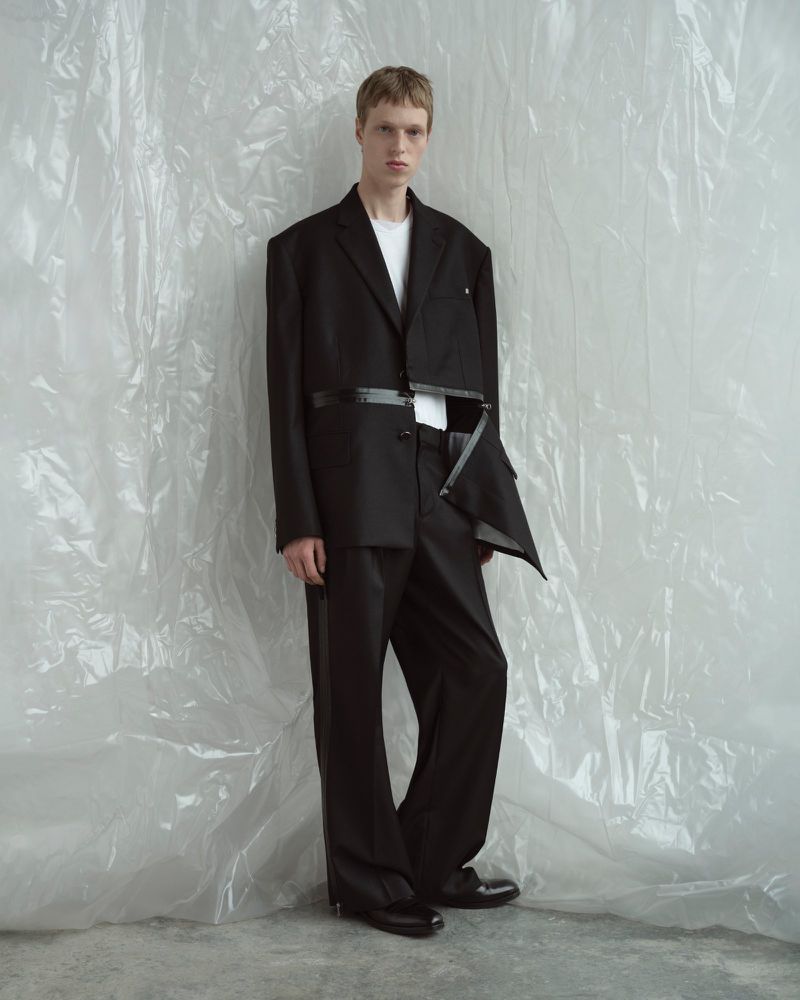 Editorial fashion shot of a young adult male model in a black deconstructed suit, posing against a textured white backdrop in studio lighting.