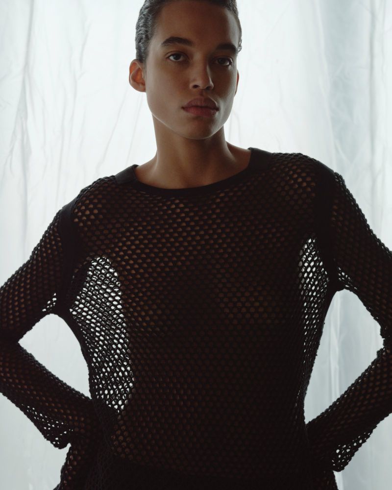 An adult with short dark hair poses in a black, net-like long-sleeved shirt against a bright white background in a studio setting.