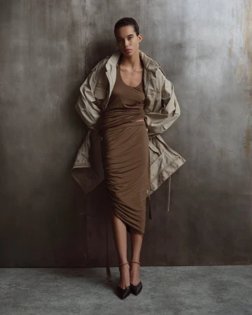 A stylish woman in a brown dress and jacket poses confidently against a neutral textured background, showcasing a fashion editorial look.