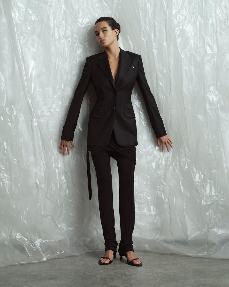 Fashion shot of a biracial model in a black blazer and black pantsuit posing against a white plastic sheet backdrop in a photo studio.