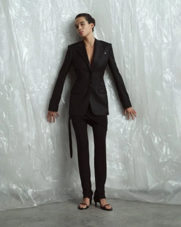 Fashion shot of a biracial model in a black blazer and black pantsuit posing against a white plastic sheet backdrop in a photo studio.