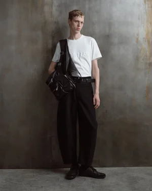 Model wearing a white t-shirt, black trousers, and a black leather bag against a distressed textured wall
