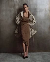 Model leaning against a textured grey wall wearing a ruched brown dress and an oversized utility jacket.