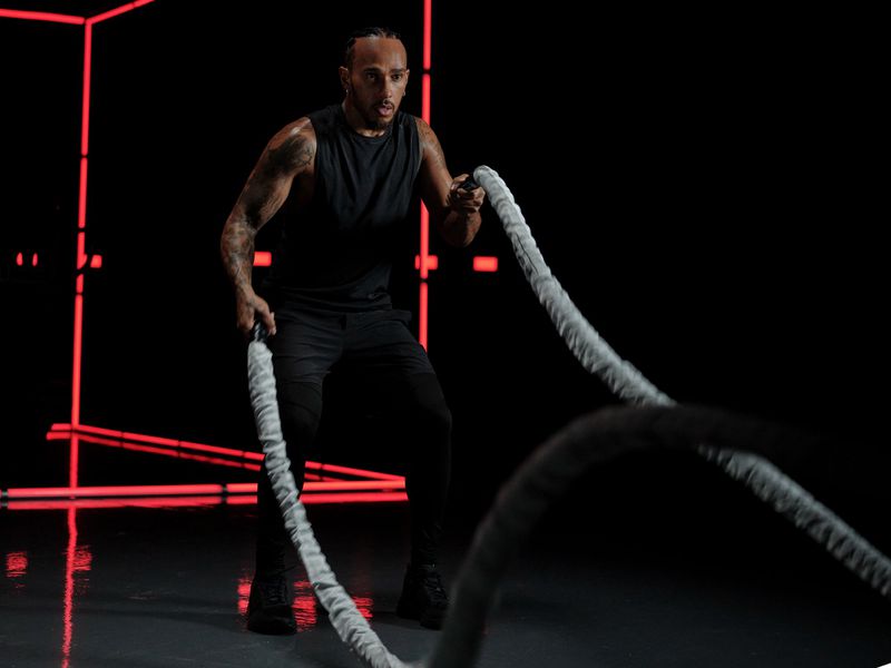 An adult male with dark hair and tattoos is exercising indoors with white ropes on a dark grey floor against a black background.