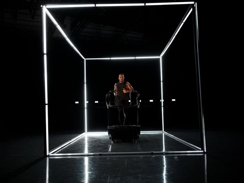 Athletic man wearing tank top jogging on a treadmill within an illuminated cube in a darkened studio, focused on fitness and technology.