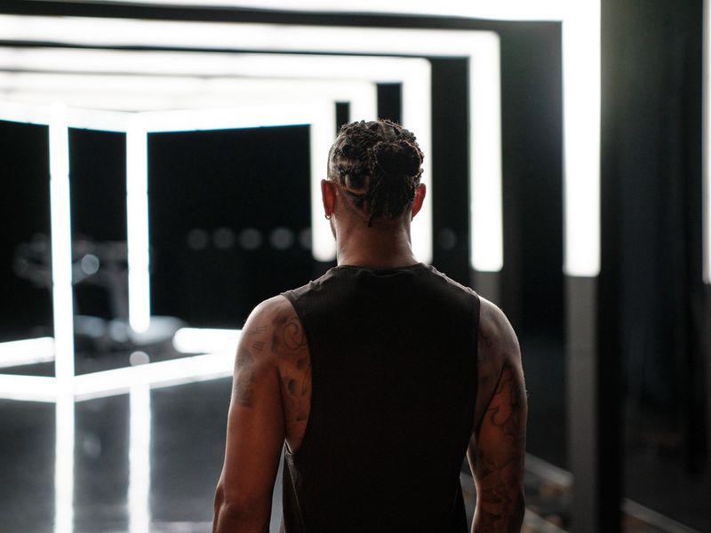 Back view of adult male with braided hair and tattoos wearing a black tank top standing in a room with illuminated cube lights.