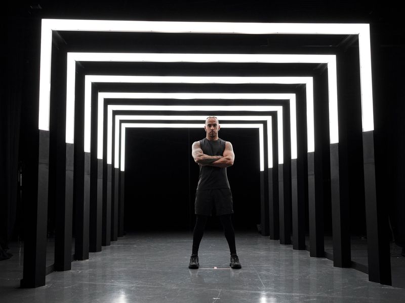 An adult male in a tank top stands confidently with crossed arms in a dark tunnel illuminated by a repeating series of white neon rectangles.