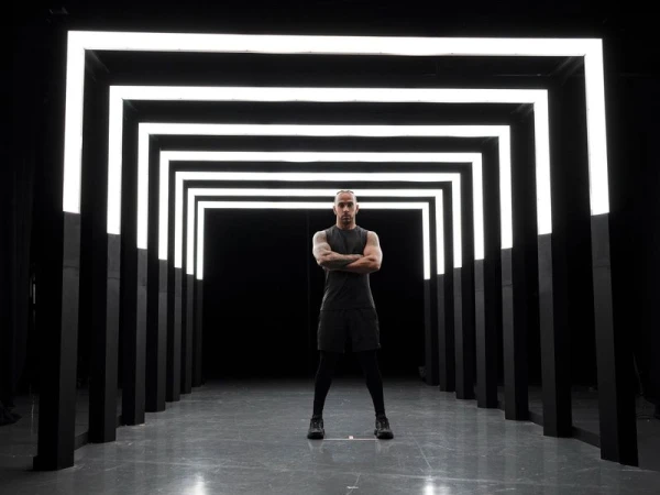 An adult male in a tank top stands confidently with crossed arms in a dark tunnel illuminated by a repeating series of white neon rectangles.