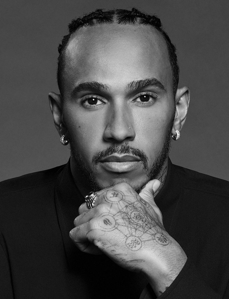 Black and white portrait of a man with braided hair wearing a collared shirt, hand jewelry, and hand tattoos against a grey backdrop.