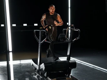 Lewis Hamilton running on a curved treadmill in a dark studio lit by vertical neon light strips