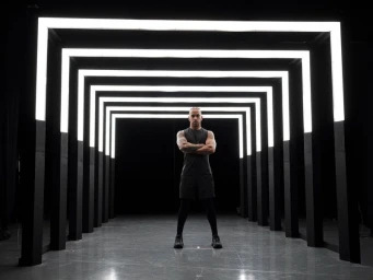 Lewis Hamilton standing with arms crossed inside a tunnel illuminated by receding square neon light frames.