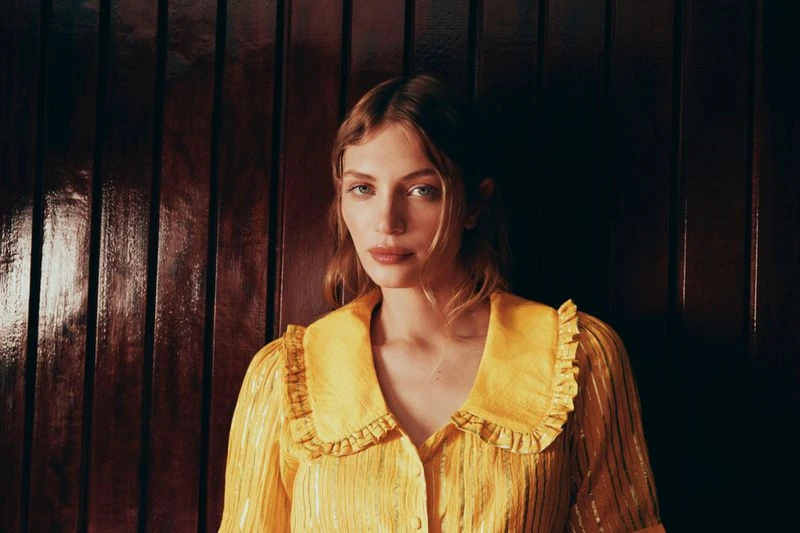 Portrait of a fair-skinned woman with blonde hair, wearing a yellow ruffle-collared blouse against a dark wood-paneled wall.