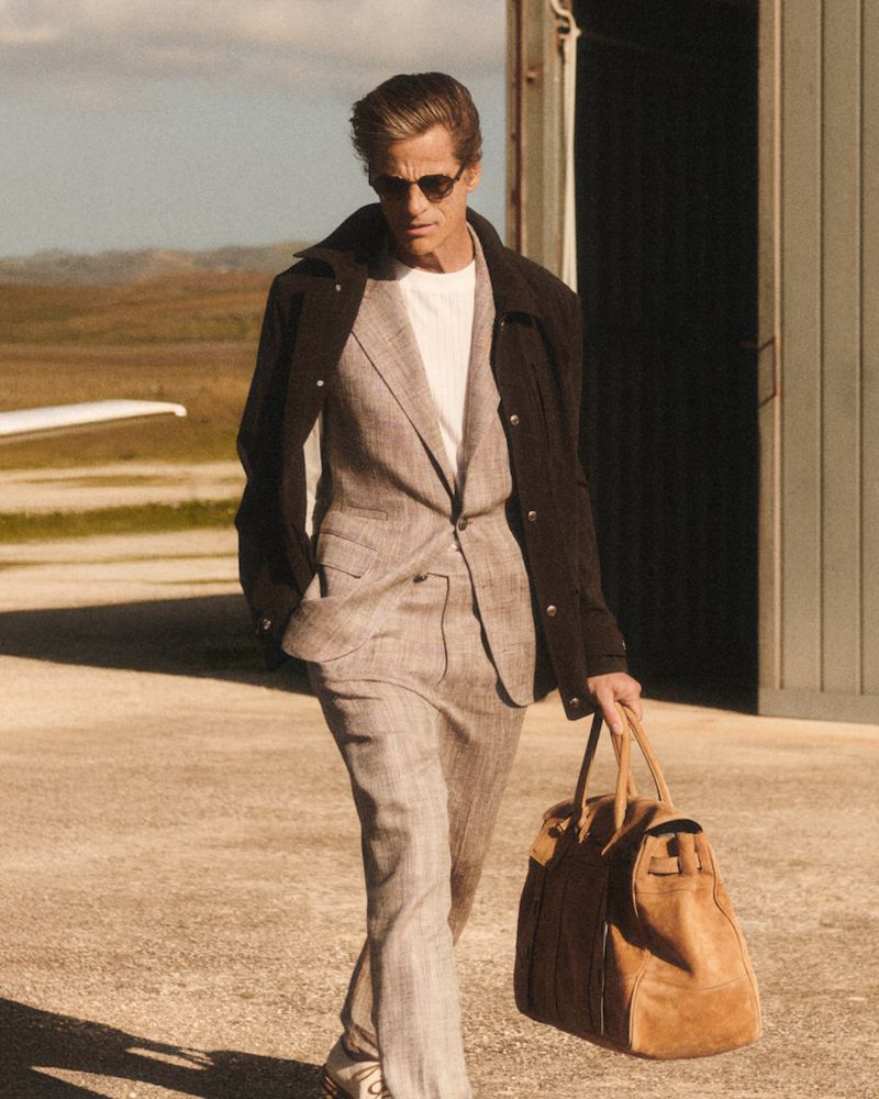 A stylish man in a suit walks on a tarmac near an aircraft hangar, carrying a duffel bag, with a small airplane in the background.