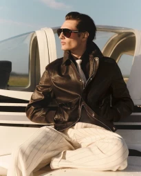A man wearing dark sunglasses, dark leather jacket, and pinstripe trousers seated outdoors near a small aircraft under a clear sky.