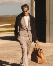 Man wearing a grey plaid suit and dark jacket holding a tan suede duffel bag walking near a hangar