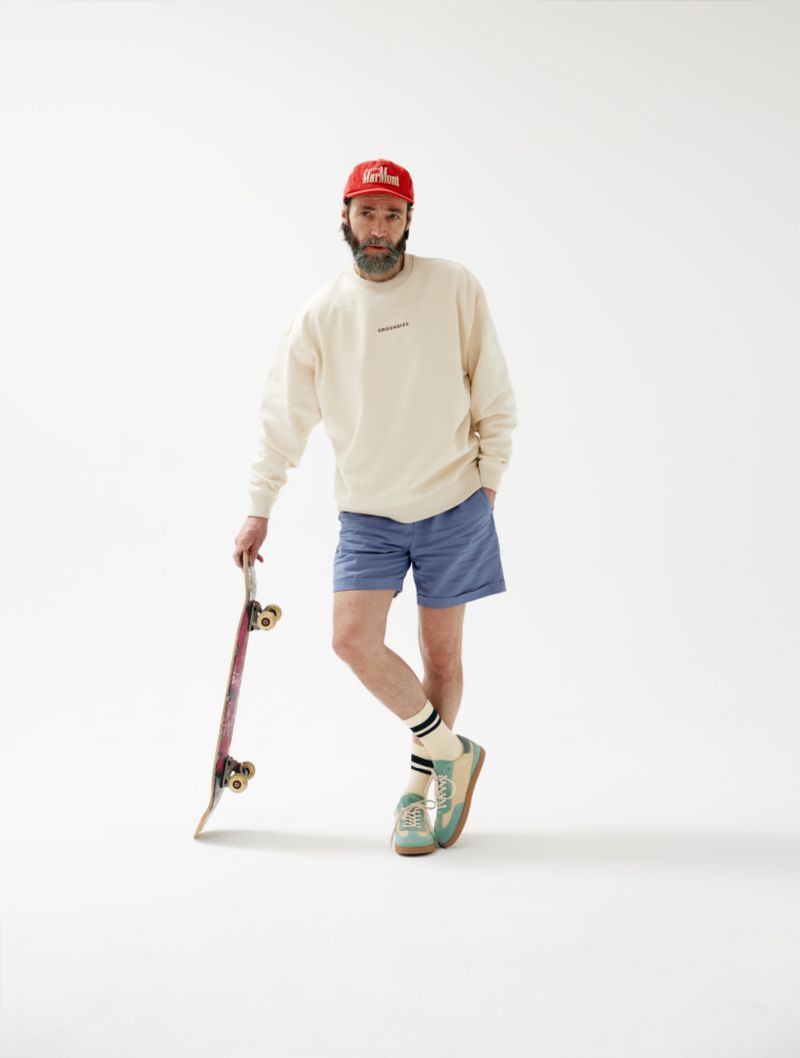 Adult male in sweatshirt, shorts, and a baseball cap, leaning on a skateboard against a plain white background in a studio shot.