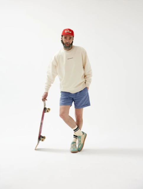 Adult male in sweatshirt, shorts, and a baseball cap, leaning on a skateboard against a plain white background in a studio shot.
