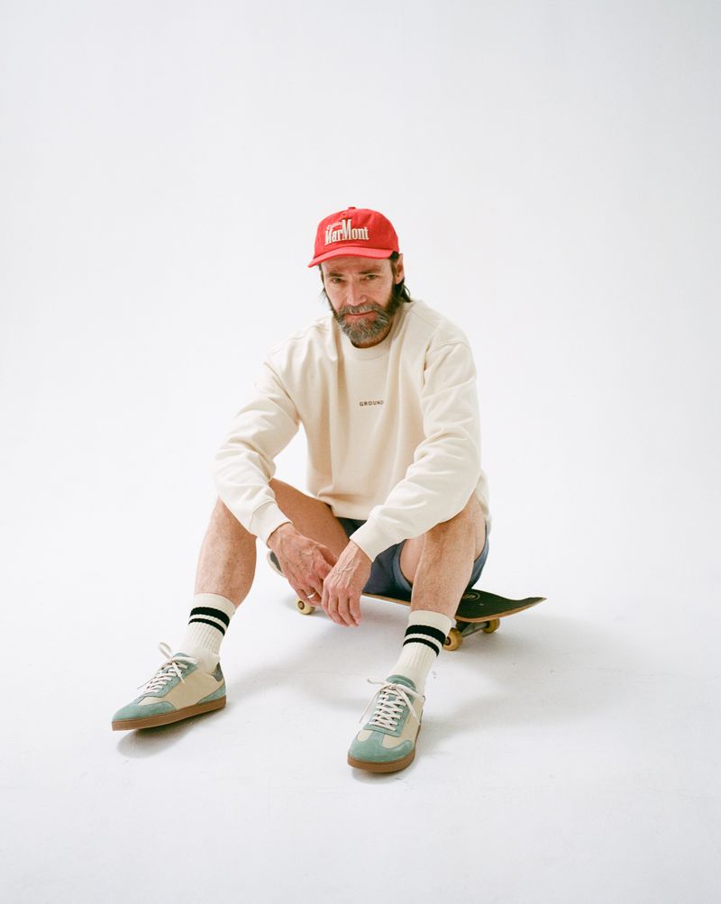 Man in sweatshirt and cap sitting on skateboard indoors in a white studio setting, wearing trainers and socks. Lifestyle shot.