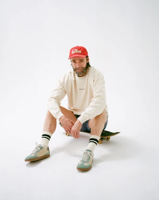 Man in sweatshirt and cap sitting on skateboard indoors in a white studio setting, wearing trainers and socks. Lifestyle shot.