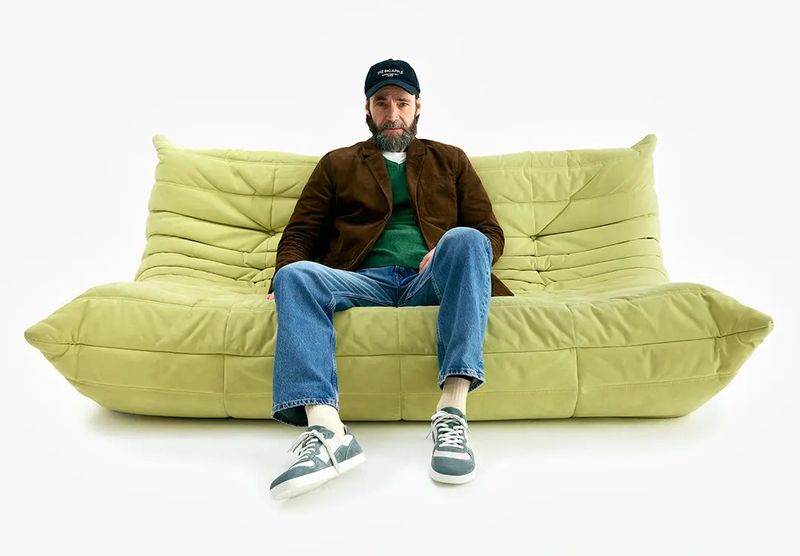 Man wearing a brown blazer, green sweater, jeans and a cap seated on a quilted light green sofa against a white background