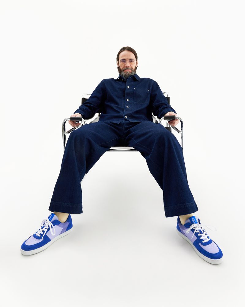 Adult male seated in metal chair in studio, wearing blue denim jumpsuit and blue and violet athletic sneakers, eye-level view.