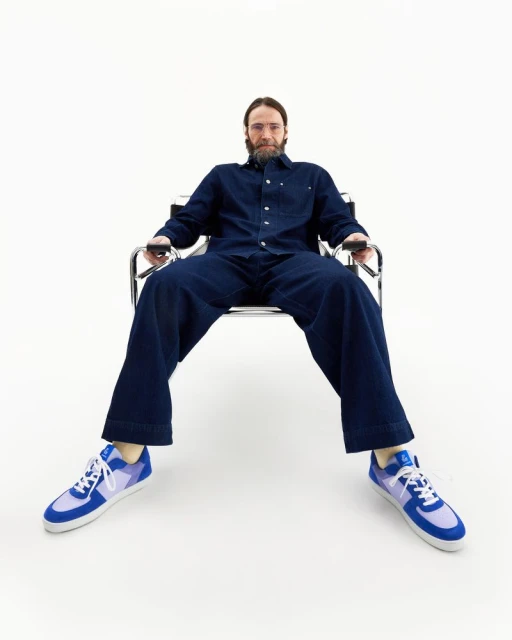 Adult male seated in metal chair in studio, wearing blue denim jumpsuit and blue and violet athletic sneakers, eye-level view.