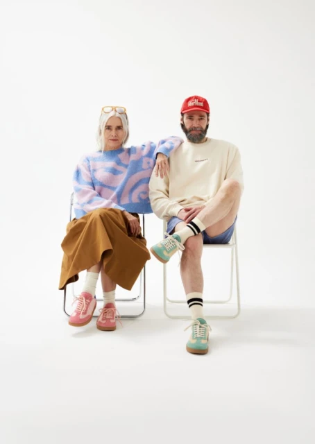 Stylish couple poses in a brightly lit photo studio, wearing trendy casual outfits, seated in chairs against a minimalist white backdrop.