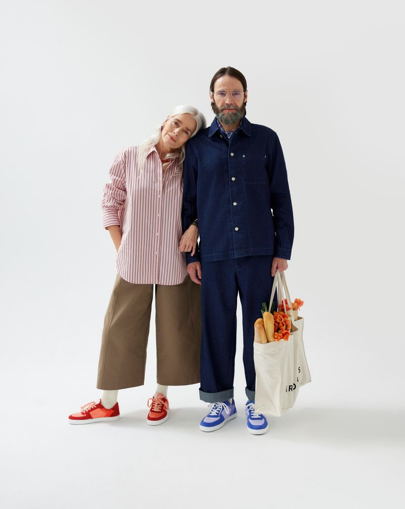An older couple poses in a photo studio in color-coordinated, casual outfits and matching sneakers while holding a tote bag with bread and flowers.