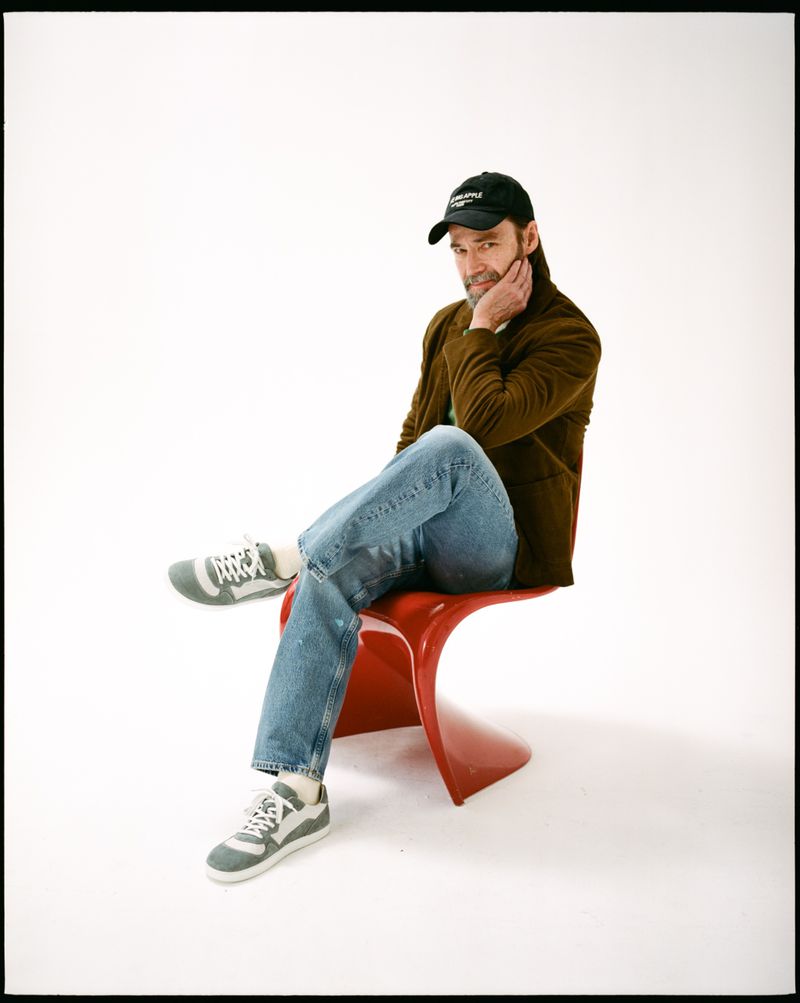 Adult male in casual attire sits on a red chair in a photo studio. He wears jeans, a brown jacket, a cap, and sneakers against a white backdrop.
