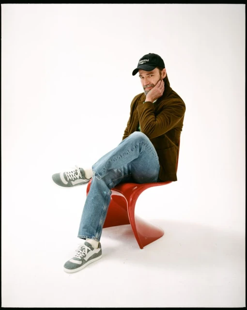 Adult male in casual attire sits on a red chair in a photo studio. He wears jeans, a brown jacket, a cap, and sneakers against a white backdrop.