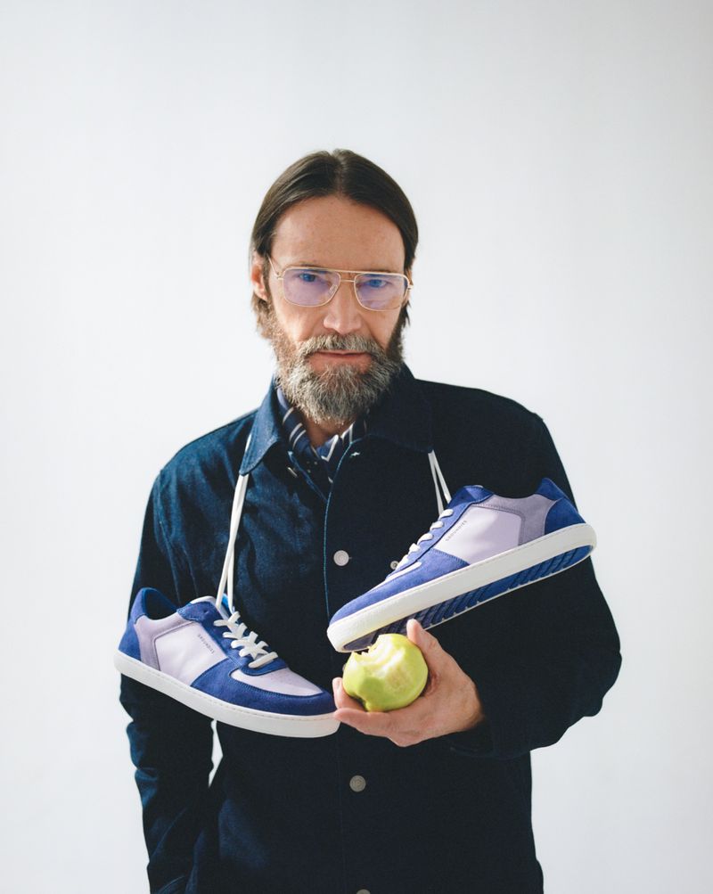 Portrait of a man with a grey beard, wearing glasses and a blue jacket, holding sneakers and an apple against a white background.