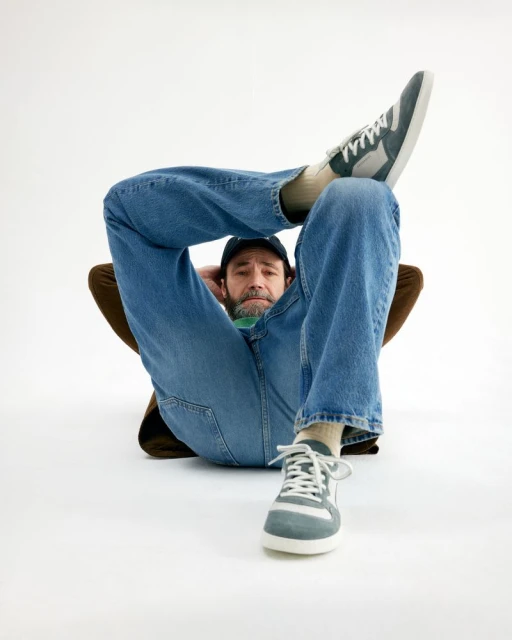 Man lying on white surface wearing jeans, blue cap, sneakers and a brown jacket. The man has a beard and dark hair.