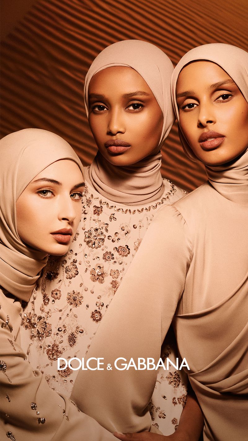 Three women wearing Dolce & Gabbana headscarves and dresses with floral embellishments pose against a tan, textured background in a portrait shot.