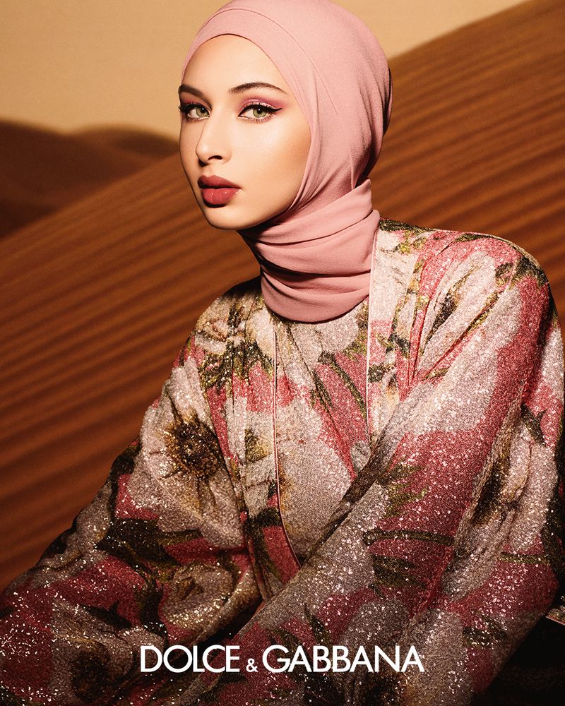 Portrait of a woman wearing a pink hijab and floral Dolce & Gabbana robe, posing in a desert landscape with soft, diffused lighting.