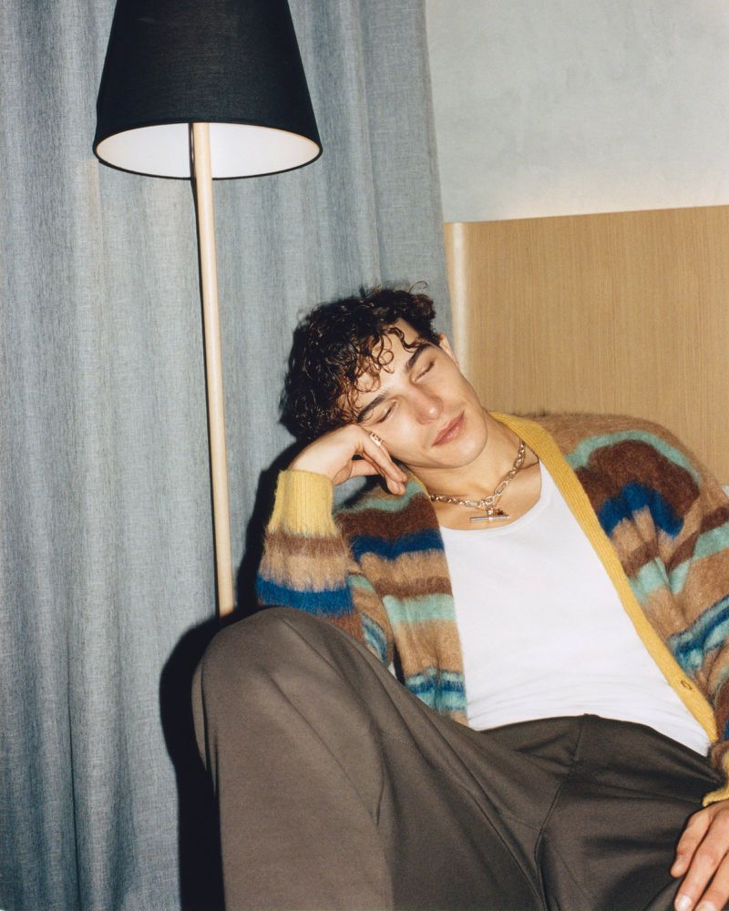 Young man with curly dark hair rests his head on his hand, wearing a striped cardigan with brown pants, next to a floor lamp and curtain.