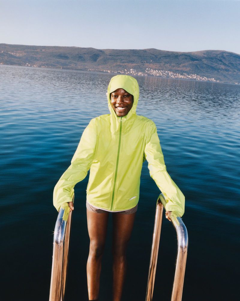 A smiling Black woman in a neon yellow jacket stands on metal steps in a lake against a background of hills and a blue sky.