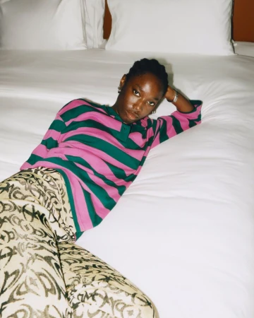 A person with dark skin and short dark hair is reclining on a white bed with pillows, wearing a striped shirt and patterned pants.
