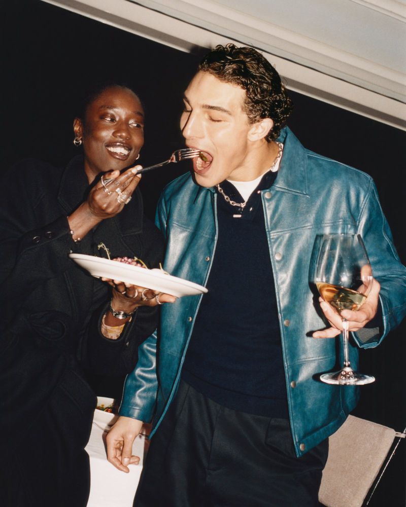 An adult woman feeds an adult man with a fork. He holds a wine glass. They both are dressed in modern fashion, indoors.