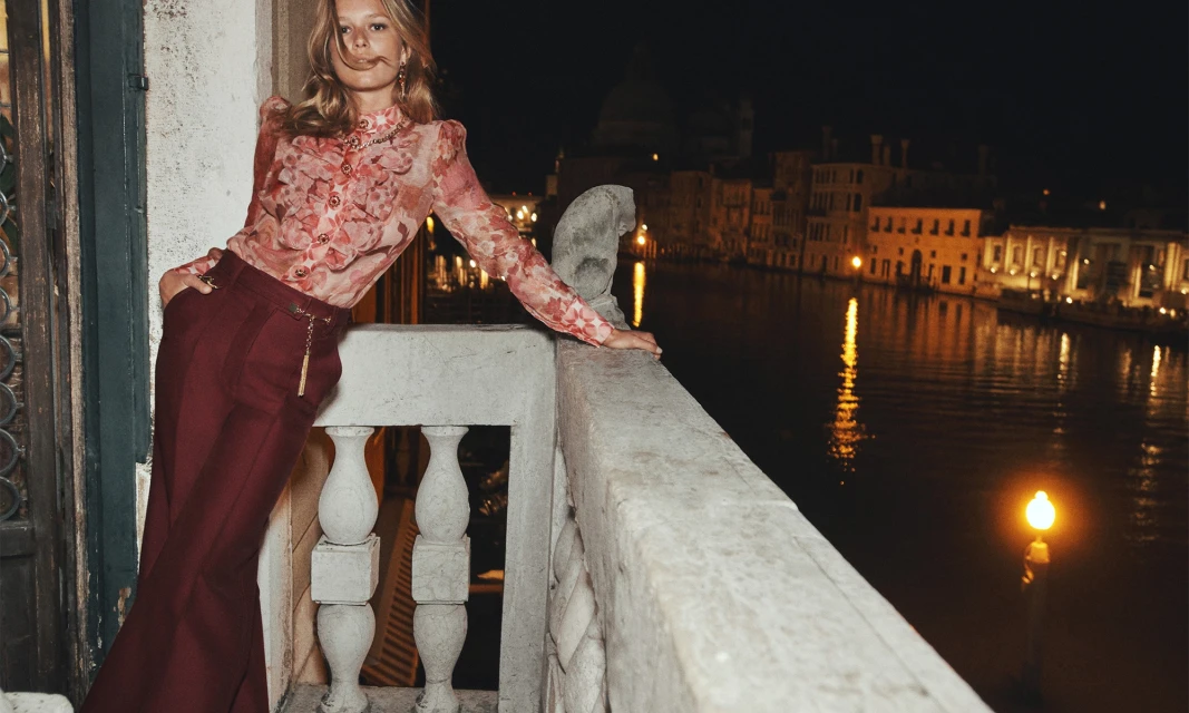 Blonde woman in a floral blouse and maroon pants poses on a balcony with a canal and illuminated buildings in the background at night.