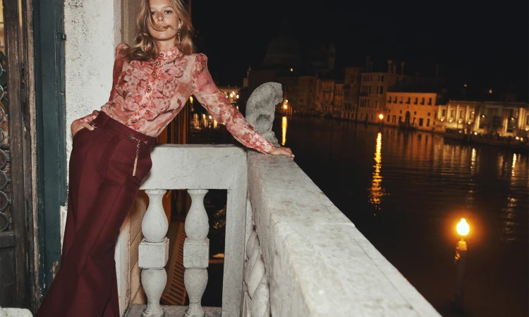 Blonde woman in a floral blouse and maroon pants poses on a balcony with a canal and illuminated buildings in the background at night.