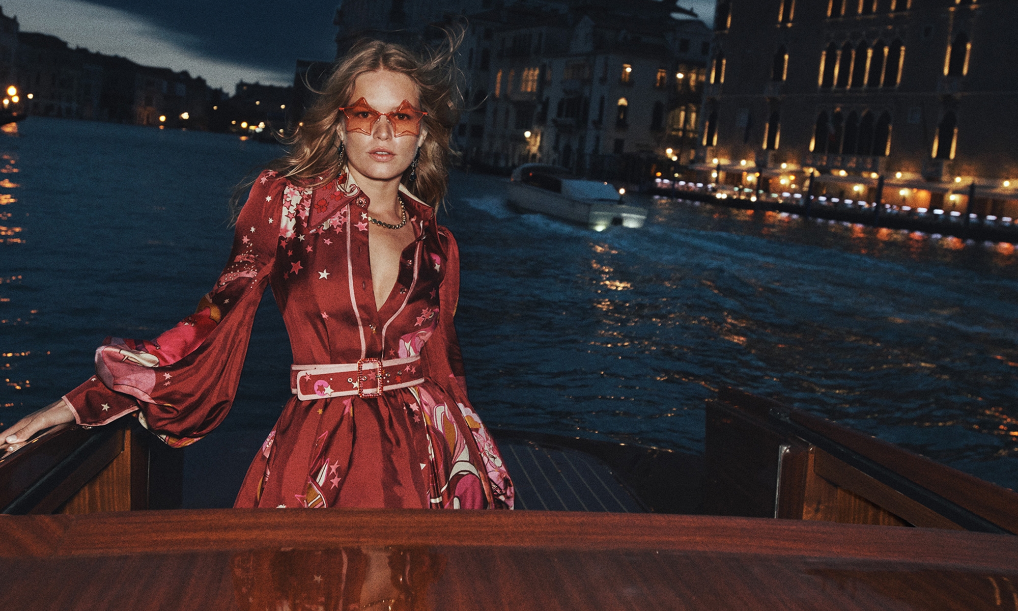 Model Anna Ewers wearing a red printed dress and tinted sunglasses on a boat at night for Zimmermann