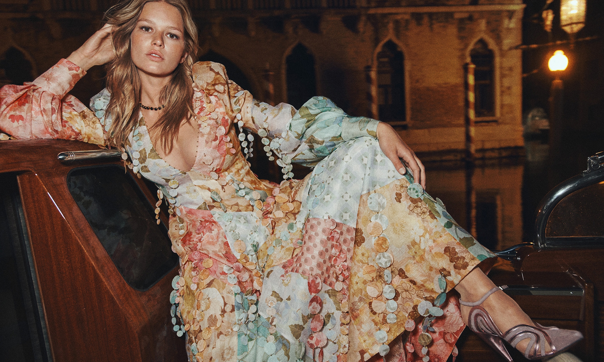 A blonde woman poses in a floral gown on a boat at night with buildings in the background under the glow of streetlights.
