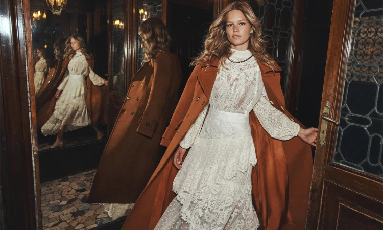 Anna Ewers in white lace dress and brown coat standing in a wood-paneled interior with mirrors.