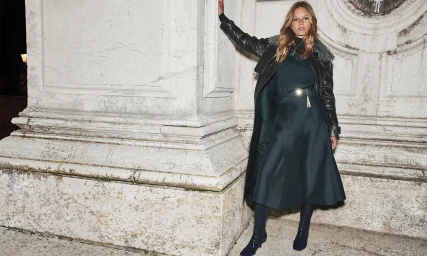 Anna Ewers leaning against a classical stone building in a dark green coat and pleated midi skirt for Zimmermann Fall RTW.